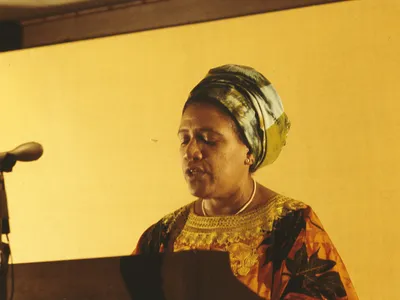 Candid color photo of Audre Lorde from the shoulders up standing at a microphone and podium.