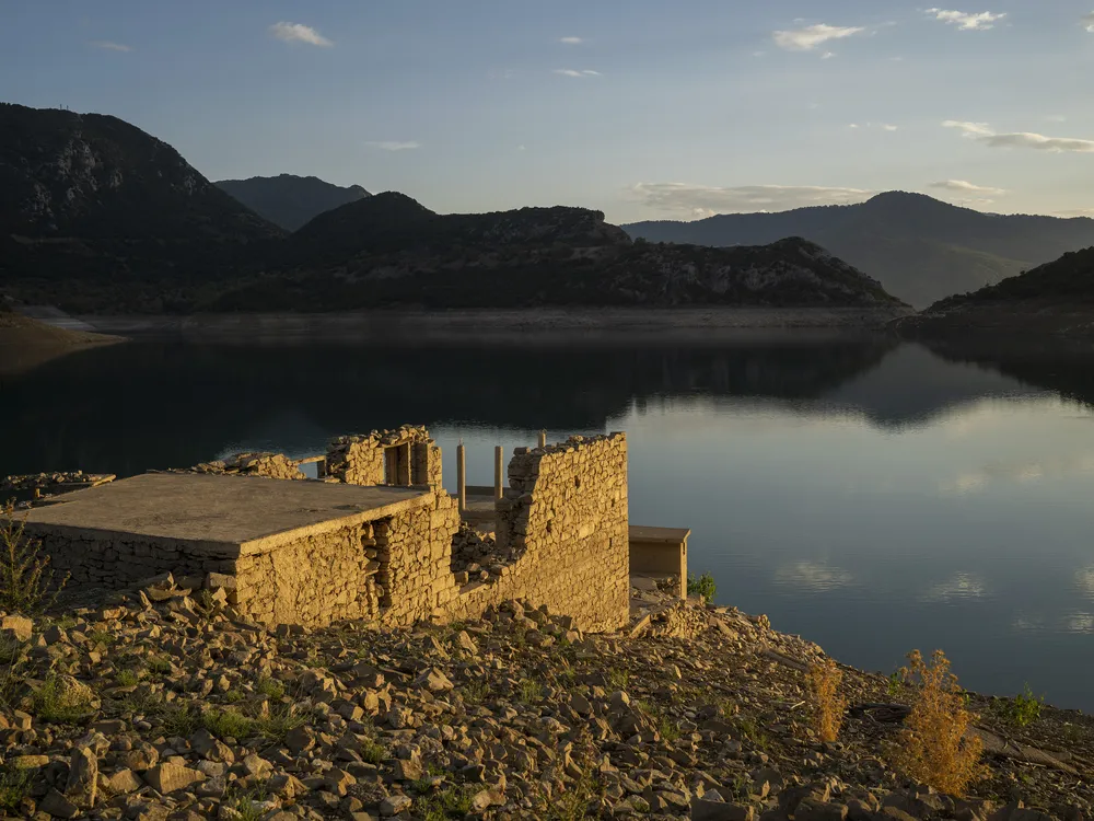 ruins of a building stand out from the side of a hill against a low lake with mountains in the background