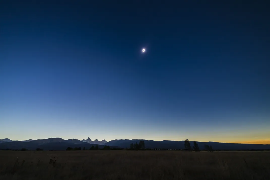 The sun during a total eclipse, seen small in the sky with the moon blocking out its center, over a dark sky with sunset colors along the horizon