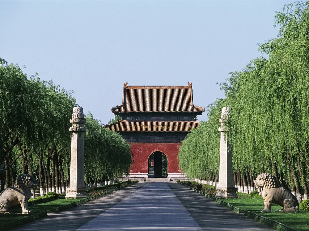 Red building at the end of a long road lined with trees and stone statues